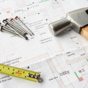 Photo of a set of building plans with a tape measure, hammer, and nails laying across the plans.